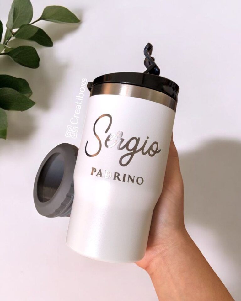 Una mano sostiene una taza de viaje blanca con el nombre Sergio Padrino impreso en letras mayúsculas y cursivas. La taza tiene una tapa negra y, al fondo, una planta con hojas verdes.