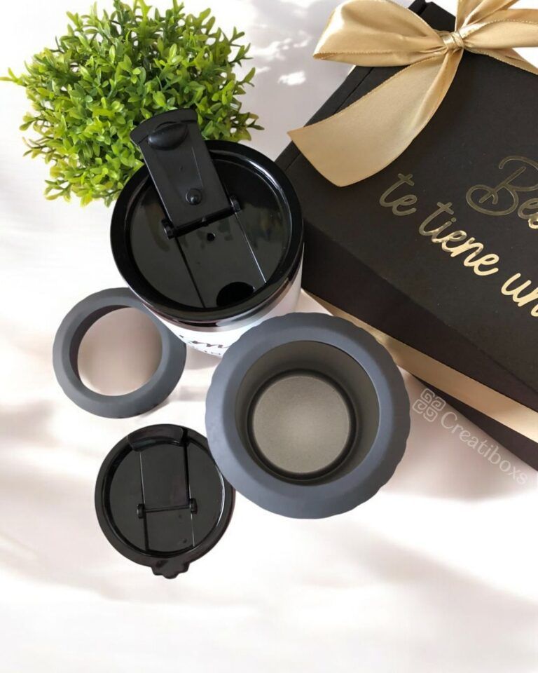 Sobre una mesa se exhibe una taza de viaje negra con tapa y piezas desmontables. Junto a ella hay una pequeña planta verde y una caja de regalo negra con una cinta dorada y letras doradas parcialmente visibles.