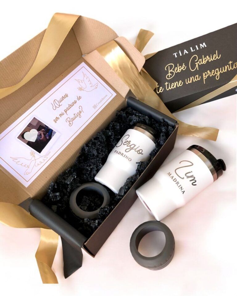Set de regalo con dos tazas térmicas blancas con la inscripción Sergio - Padrino y Lia - Madrina. La caja incluye una tarjeta con una foto y un texto, parcialmente visible, y material de embalaje protector negro. También se incluye un sobre decorado.