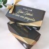 Dos cajas de regalo negras con cintas doradas y texto. La caja superior dice Tía Lim Bebé Gabriel te tiene una pregunta. La caja inferior también tiene una cinta dorada. Se ve una pequeña planta verde en la esquina.