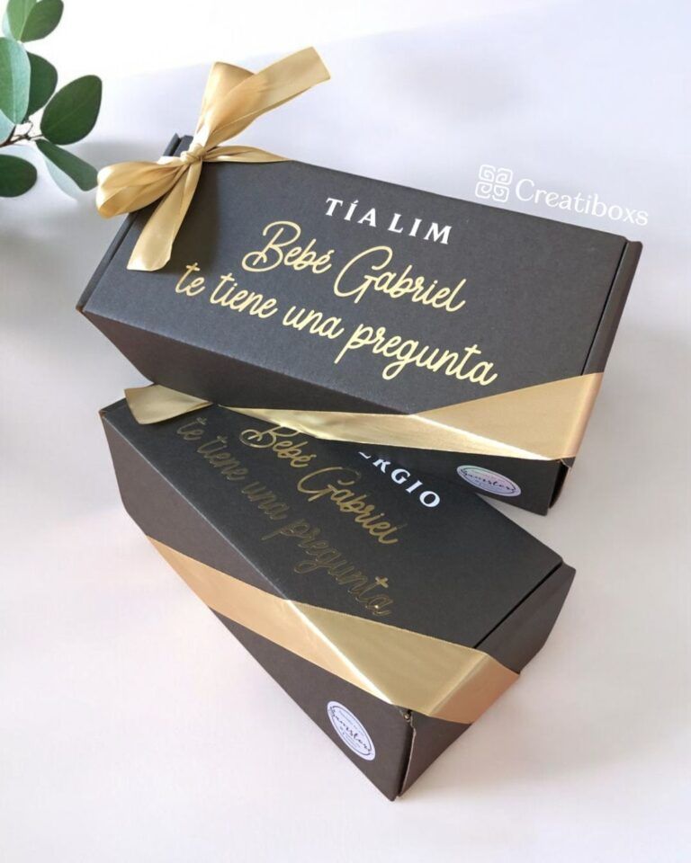 Dos cajas de regalo negras con cintas doradas y texto. La caja superior dice Tía Lim Bebé Gabriel te tiene una pregunta. La caja inferior también tiene una cinta dorada. Se ve una pequeña planta verde en la esquina.