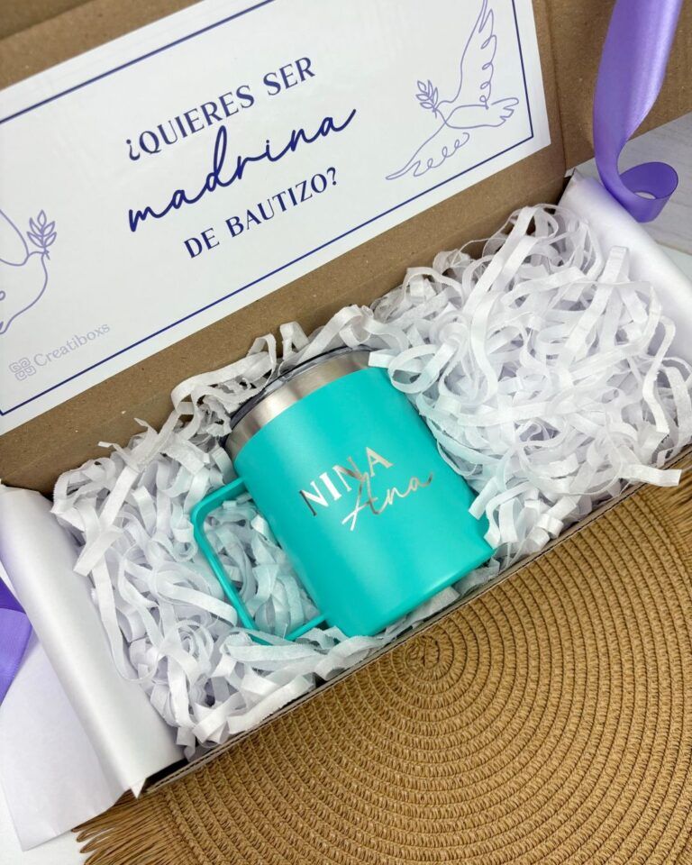 Una taza verde azulado con el texto Nina Ana se coloca dentro de una caja de regalo llena de papel arrugado blanco. Sobre la taza, un cartel dice ¿Quieres ser madrina de bautizo?. La caja está sobre una estera tejida. Hay cintas moradas a cada lado de la caja.