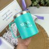 Una mano sostiene una taza turquesa con el texto Nina Ana impreso. La taza está dibujada sobre una superficie marrón. En el fondo, hay una caja decorativa con una tarjeta y cintas moradas.