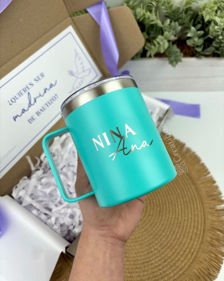 Una mano sostiene una taza turquesa con el texto Nina Ana impreso. La taza está dibujada sobre una superficie marrón. En el fondo, hay una caja decorativa con una tarjeta y cintas moradas.