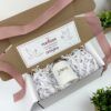 Una caja de regalo con una taza blanca en su interior, rodeada de papel triturado blanco. La taza tiene impreso el nombre Lorena. La tapa de la caja tiene un mensaje en español con ilustraciones de palomas y un corazón. Una cinta de raso rosa bordea la caja.