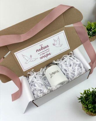 Una caja de regalo con una taza blanca en su interior, rodeada de papel triturado blanco. La taza tiene impreso el nombre Lorena. La tapa de la caja tiene un mensaje en español con ilustraciones de palomas y un corazón. Una cinta de raso rosa bordea la caja.