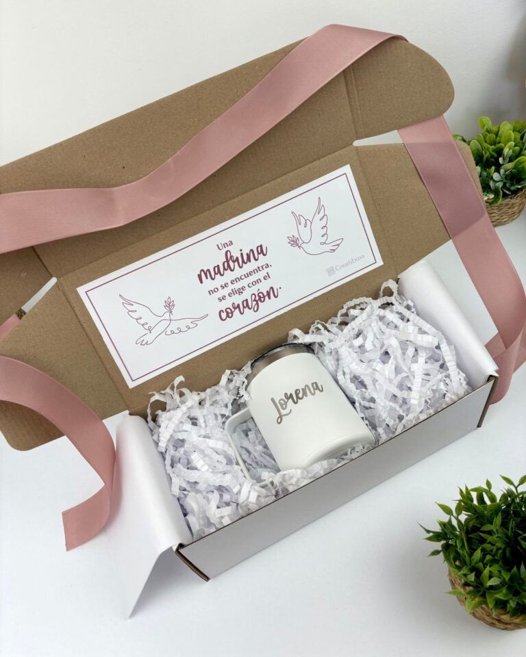 Una caja de regalo con una taza blanca en su interior, rodeada de papel triturado blanco. La taza tiene impreso el nombre Lorena. La tapa de la caja tiene un mensaje en español con ilustraciones de palomas y un corazón. Una cinta de raso rosa bordea la caja.