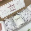 El paquete incluye una taza blanca con la palabra Lorena impresa, rodeada de papel arrugado blanco. Encima de la taza hay una tarjeta con texto rosa e ilustraciones de palomas que dice Una madrina no se encuentra, se elige con el corazón.