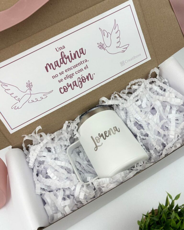 El paquete incluye una taza blanca con la palabra Lorena impresa, rodeada de papel arrugado blanco. Encima de la taza hay una tarjeta con texto rosa e ilustraciones de palomas que dice Una madrina no se encuentra, se elige con el corazón.