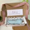 Una caja de cartón abierta con una cinta rosa contiene un vaso azul claro con la etiqueta Perla en texto dorado. El vaso está rodeado de papel triturado. La tapa interior muestra una tarjeta con texto en español sobre ser madrina para la comunión y la confirmación.