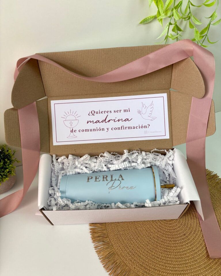 Una caja de cartón abierta con una cinta rosa contiene un vaso azul claro con la etiqueta Perla en texto dorado. El vaso está rodeado de papel triturado. La tapa interior muestra una tarjeta con texto en español sobre ser madrina para la comunión y la confirmación.