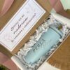 Un vaso azul claro con la palabra PERLA impresa se encuentra dentro de una caja de regalo llena de papel arrugado blanco. Una tarjeta dice ¿Quieres ser mi madrina de comunión y confirmación? junto con imágenes de un pájaro y un cáliz.