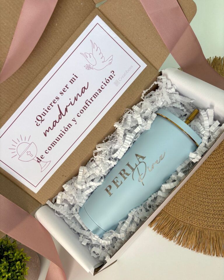 Un vaso azul claro con la palabra PERLA impresa se encuentra dentro de una caja de regalo llena de papel arrugado blanco. Una tarjeta dice ¿Quieres ser mi madrina de comunión y confirmación? junto con imágenes de un pájaro y un cáliz.