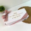Una caja de regalo blanca con un lazo de cinta rosa, colocada sobre una estera tejida. El texto en la caja dice Tía hay algo que quiero preguntarte. Una pequeña planta en maceta se ve en el fondo sobre una superficie blanca.