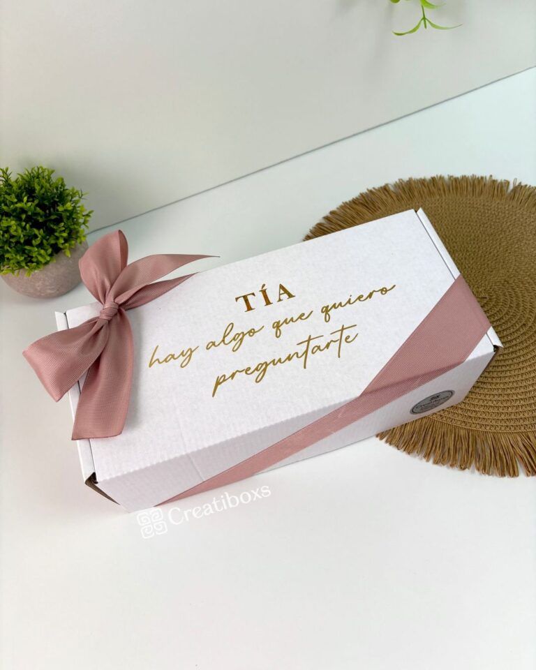 Una caja de regalo blanca con un lazo de cinta rosa, colocada sobre una estera tejida. El texto en la caja dice Tía hay algo que quiero preguntarte. Una pequeña planta en maceta se ve en el fondo sobre una superficie blanca.