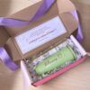 Una caja de regalo abierta contiene una taza verde con el nombre Blanca y un corazón, una pajita de metal y un limpiador de pajitas. Se incluye una tarjeta con elementos decorativos y texto en español. La caja está forrada con papel arrugado y tiene una cinta morada.