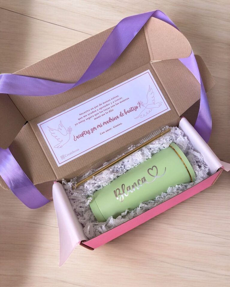 Una caja de regalo abierta contiene una taza verde con el nombre Blanca y un corazón, una pajita de metal y un limpiador de pajitas. Se incluye una tarjeta con elementos decorativos y texto en español. La caja está forrada con papel arrugado y tiene una cinta morada.
