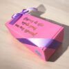Sobre una superficie de madera clara se coloca una caja de regalo rosa con una cinta violeta. La caja tiene letras doradas que dicen: Después de este regalo serás aún más especial.