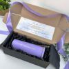 Una caja de regalo rectangular de cartón con un lazo violeta contiene un vaso térmico violeta con la inscripción Andrea en letras doradas. Se incluye una nota en la tapa y dos pequeñas plantas verdes en el fondo.