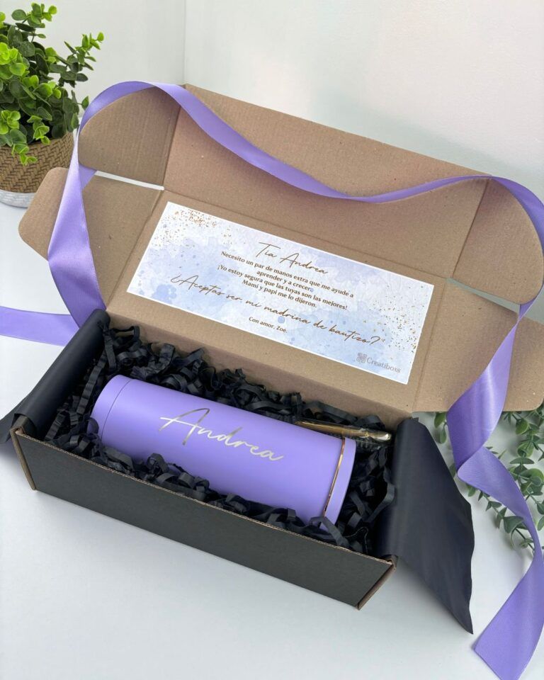Una caja de regalo rectangular de cartón con un lazo violeta contiene un vaso térmico violeta con la inscripción Andrea en letras doradas. Se incluye una nota en la tapa y dos pequeñas plantas verdes en el fondo.