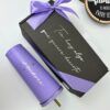 Un vaso morado con el nombre Andrea junto a una caja de regalo negra adornada con un lazo morado y un mensaje en español. En el fondo se ve parcialmente una decoración redonda de madera con una cita inspiradora.