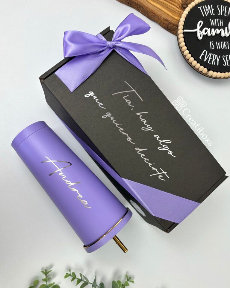 Un vaso morado con el nombre Andrea junto a una caja de regalo negra adornada con un lazo morado y un mensaje en español. En el fondo se ve parcialmente una decoración redonda de madera con una cita inspiradora.