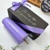 Un vaso cilíndrico morado con el nombre Andrea en cursiva al lado de una caja de regalo negra atada con una cinta morada. La caja tiene la inscripción Te hay algo que quiero decirte en cursiva blanca. En el fondo hay pequeñas hojas verdes y accesorios de madera.