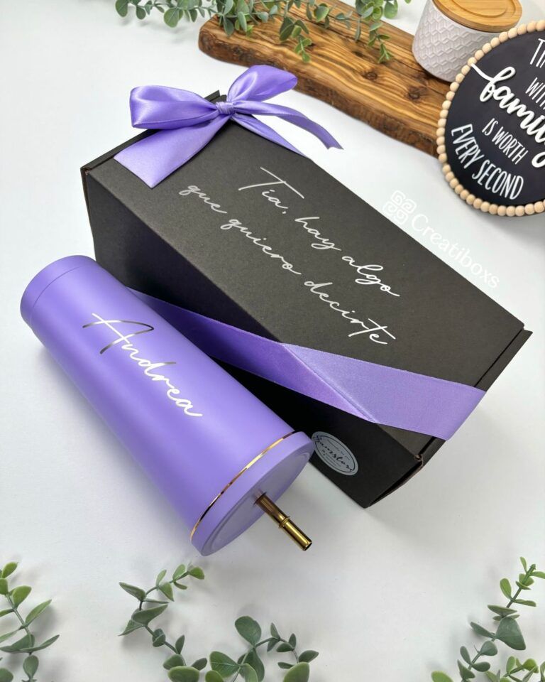 Un vaso cilíndrico morado con el nombre Andrea en cursiva al lado de una caja de regalo negra atada con una cinta morada. La caja tiene la inscripción Te hay algo que quiero decirte en cursiva blanca. En el fondo hay pequeñas hojas verdes y accesorios de madera.