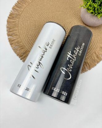 Sobre una estera tejida se encuentran dos vasos de acero inoxidable. El vaso de la izquierda es blanco y lleva la etiqueta Alejandra Bride y el vaso de la derecha es negro y lleva la etiqueta Jonathan Groom. Ambos llevan la fecha 02.12.2023. Una pequeña planta es parcialmente visible sobre ellos.