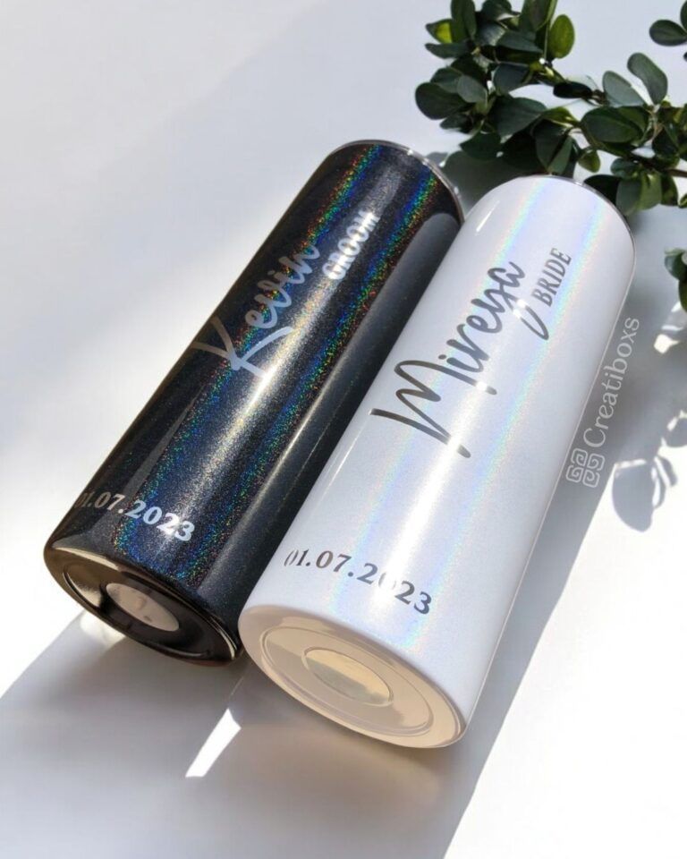 Sobre una superficie yacen dos botellas cilíndricas con diseños holográficos. La botella negra lleva la etiqueta Kevin y la blanca Mireya, con la palabra Bride (novia). Ambas tienen impresa la fecha 01.07.2023. Cerca hay una ramita de vegetación.