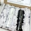Una caja de regalo contiene dos vasos personalizados, uno plateado con la etiqueta Bride (Novia) y el nombre Alejandra y el otro negro con la etiqueta Groom (Novio) y el nombre Jonathan. Ambos tienen la fecha 02.12.2023. La caja está forrada con trozos de papel blanco.