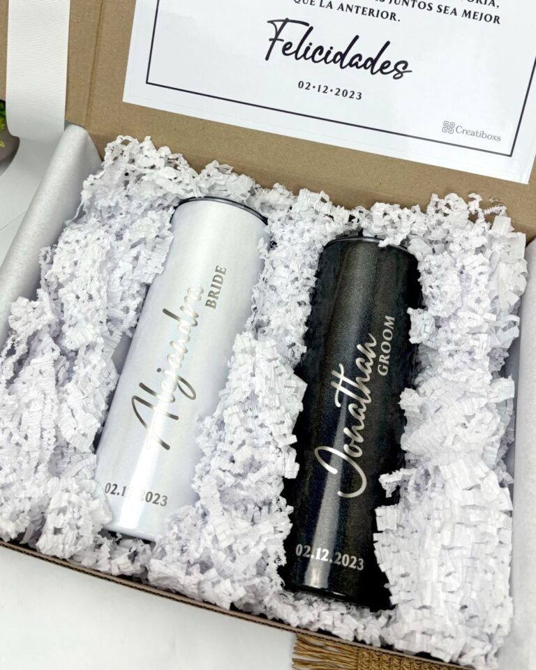 Una caja de regalo contiene dos vasos personalizados, uno plateado con la etiqueta Bride (Novia) y el nombre Alejandra y el otro negro con la etiqueta Groom (Novio) y el nombre Jonathan. Ambos tienen la fecha 02.12.2023. La caja está forrada con trozos de papel blanco.
