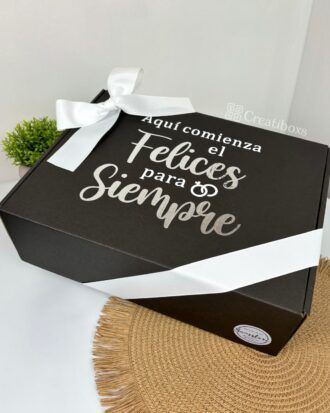 Una caja de regalo negra con el texto blanco Aquí comienza el feliz para siempre reposa sobre una estera tejida. La caja está atada con una cinta blanca. En el fondo se ve una pequeña planta verde en maceta.