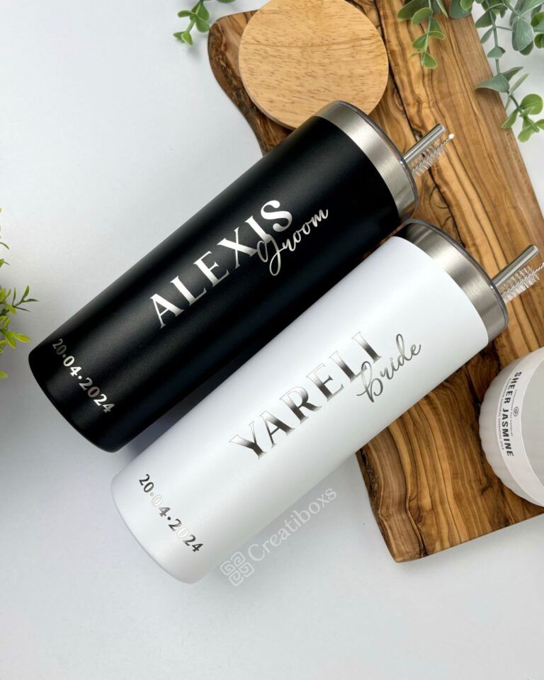 Sobre una tabla de madera con hojas verdes se colocan dos vasos de acero inoxidable. Uno es negro y lleva la inscripción Alexis Groom y la fecha 20.04.2024, mientras que el otro es blanco y lleva la inscripción Yareli Bride y la misma fecha.