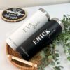 Dos vasos personalizados, uno blanco con la etiqueta Eva Bride y el otro negro con la etiqueta Erick Groom, se colocan en una canasta tejida con hojas verdes. Cada vaso tiene la fecha 23.03.2024. Un cartel dice El tiempo pasado con la familia vale cada segundo.