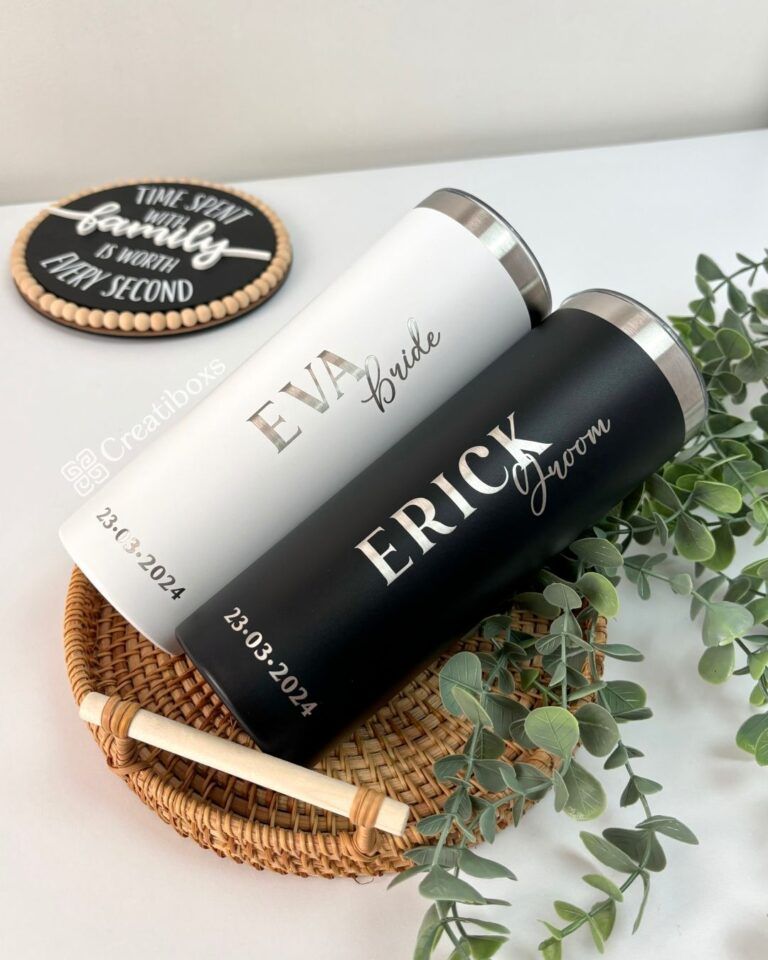 Dos vasos personalizados, uno blanco con la etiqueta Eva Bride y el otro negro con la etiqueta Erick Groom, se colocan en una canasta tejida con hojas verdes. Cada vaso tiene la fecha 23.03.2024. Un cartel dice El tiempo pasado con la familia vale cada segundo.