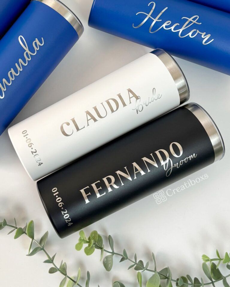 Se muestran cuatro vasos personalizados. Dos vasos azules con nombres en cursiva, un vaso blanco con la etiqueta Claudia Bride y un vaso negro con la etiqueta Fernando Groom, ambos con fecha del 01.06.2024. Se ven hojas de eucalipto en las esquinas inferiores.