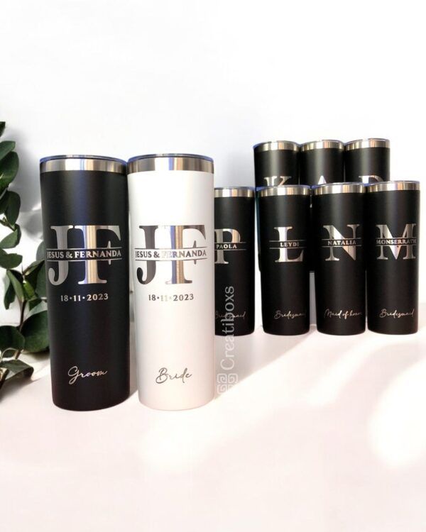 Un juego de vasos personalizados con iniciales y nombres. Dos en el frente tienen la etiqueta Novio y Novia con la fecha de la boda, mientras que los otros en la parte posterior tienen la etiqueta de la fiesta de bodas. Se ve una pequeña planta en el costado.