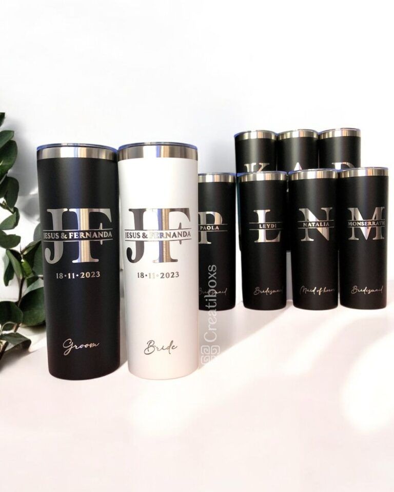 Un juego de vasos personalizados con iniciales y nombres. Dos en el frente tienen la etiqueta Novio y Novia con la fecha de la boda, mientras que los otros en la parte posterior tienen la etiqueta de la fiesta de bodas. Se ve una pequeña planta en el costado.