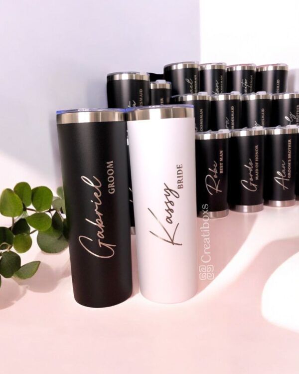 En primer plano se ven dos vasos personalizados con la inscripción Novio y Novia. El vaso del novio es negro y el de la novia es blanco. En el fondo se ven varios vasos más apilados. A la izquierda hay una pequeña planta.