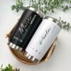 Dos tazas de viaje personalizadas, una con la etiqueta negra Moisés novio y la otra con la etiqueta blanca Paola novia, se colocan en una canasta tejida. La fecha en ambas tazas es 12.04.2024. Alrededor de la canasta hay hojas verdes.
