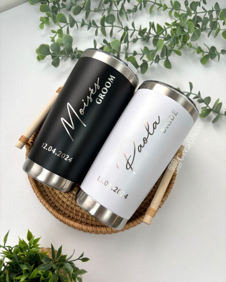 Dos tazas de viaje personalizadas, una con la etiqueta negra Moisés novio y la otra con la etiqueta blanca Paola novia, se colocan en una canasta tejida. La fecha en ambas tazas es 12.04.2024. Alrededor de la canasta hay hojas verdes.
