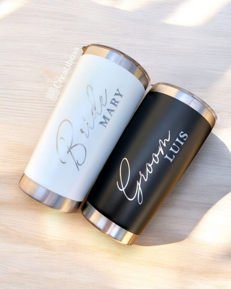 Dos vasos de viaje sobre una superficie de madera clara. El vaso blanco tiene la inscripción Bride Mary en una elegante escritura y el vaso negro tiene la inscripción Groom Luis en una escritura similar. Ambos tienen tapas y bases de acero inoxidable.