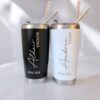 Se muestran dos vasos de acero inoxidable, uno de color negro con la etiqueta Aldair Groom y otro de color blanco con la etiqueta Andrea Bride, ambos con la fecha 20.05.2023. Cada vaso tiene una pajita de metal y un cepillo de limpieza.