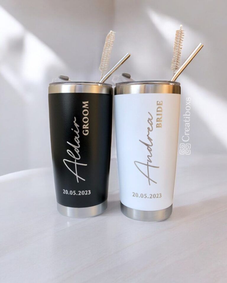 Se muestran dos vasos de acero inoxidable, uno de color negro con la etiqueta Aldair Groom y otro de color blanco con la etiqueta Andrea Bride, ambos con la fecha 20.05.2023. Cada vaso tiene una pajita de metal y un cepillo de limpieza.