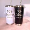 Dos vasos de acero inoxidable con pajitas, uno blanco y otro negro. El vaso blanco tiene escrito Sra. Iracema y el vaso negro Sr. Edwin. Ambos tienen escrita la fecha 30-09-2023. Los vasos están sobre una superficie de madera.