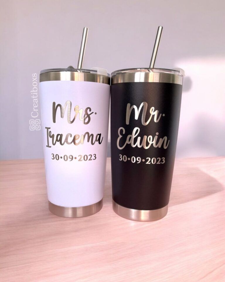 Dos vasos de acero inoxidable con pajitas, uno blanco y otro negro. El vaso blanco tiene escrito Sra. Iracema y el vaso negro Sr. Edwin. Ambos tienen escrita la fecha 30-09-2023. Los vasos están sobre una superficie de madera.