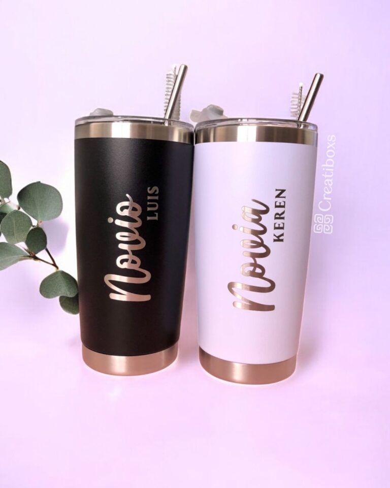 Se muestran dos vasos térmicos sobre un fondo rosa claro. El vaso negro tiene la etiqueta Novio Luis y el vaso blanco tiene la etiqueta Novia Keren. Ambos tienen pajitas de metal y bases de color dorado. Una rama verde decora el lado izquierdo.