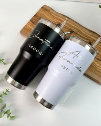 Dos vasos de acero inoxidable sobre una tabla de madera. El vaso negro tiene impresos en blanco Jonathan y Groom. El vaso blanco tiene impresos en dorado Ada Fernanda y Bride. Se ve una pequeña planta en la esquina.