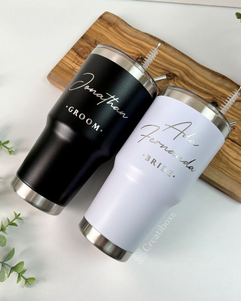 Dos vasos de acero inoxidable sobre una tabla de madera. El vaso negro tiene impresos en blanco Jonathan y Groom. El vaso blanco tiene impresos en dorado Ada Fernanda y Bride. Se ve una pequeña planta en la esquina.